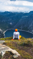Fototapeta na wymiar A woman hiker outdoor in orange and white sits on a grassy cliff face in a relaxed pose and looks at a lake far below in a deep mountain gorge. Clouds and distant snowy mountains. Summer in Norway.