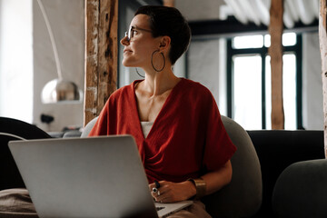 Young smiling woman working on laptop while sitting on sofa indoors