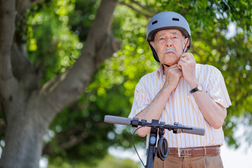 uomo anziano si sistema il  caschetto vicino al suo monopattino elettrico in una pista ciclabile vicino a un parco cittadino . - obrazy, fototapety, plakaty