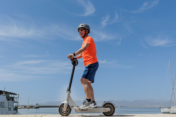 Uomo anziano con una maglia arancione e caschetto protettivo , corre sul suo monopattino elettrico  di fronte al porto turistico ., isolato su sfondo cielo . 