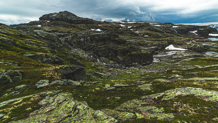 Typical Norwegian mountain landscape. Bare gray rocks covered with green moss are smoothed by the passage of a glacier, a mountain stream flows, it is raining from a gray cloudy sky, some snow.