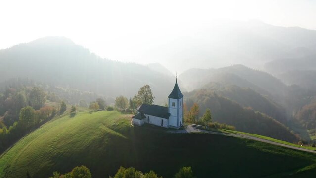 Aerial view of small church on the hill and low clouds at sunrise in autumn. Slovenia. Top view from drone of beautiful chapel on mountain in fog, green meadows, trees, sky at dawn in fall. Landscape