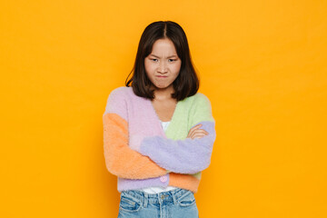 Upset asian girl standing with arms folded isolated over yellow background