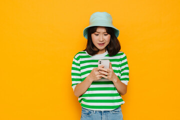 Young confused asian girl using mobile phone isolated over yellow background