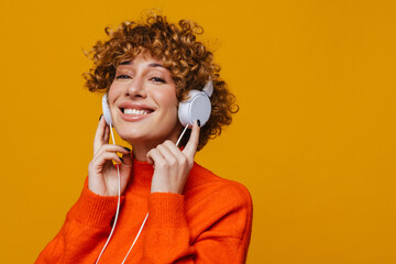 Middle-aged ginger woman listening music with headphones isolated over yellow background