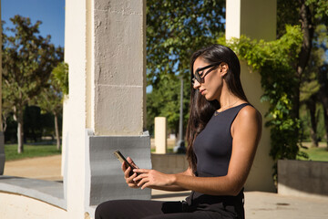 Young woman, brunette, slender, dressed in black and wearing sunglasses, sitting on a bench consulting social networks on her cell phone. Concept beauty, fashion, applications, internet, smartphone.