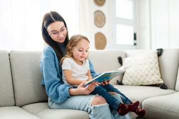 happy kid sitting on sofa with babysitter teen holding book - obrazy, fototapety, plakaty