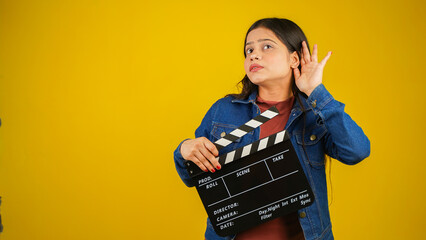 Beautiful young Asian Indian woman standing holding clapperboard, trying to listen something,...