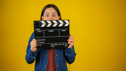 Beautiful young Asian Indian woman standing holding clapperboard shocked, clapper board used in...