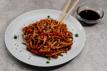 Spicy udon with chicken on a white plate with soy sauce in a glass bowl on a gray background. Side view