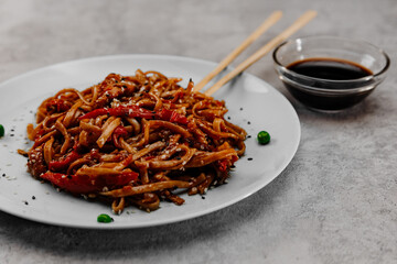 Spicy udon with chicken on a white plate with soy sauce in a glass bowl on a gray background. Side...