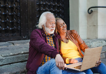 Multiracial senior couple having fun using computer laptop while sitting on stairs outdoor