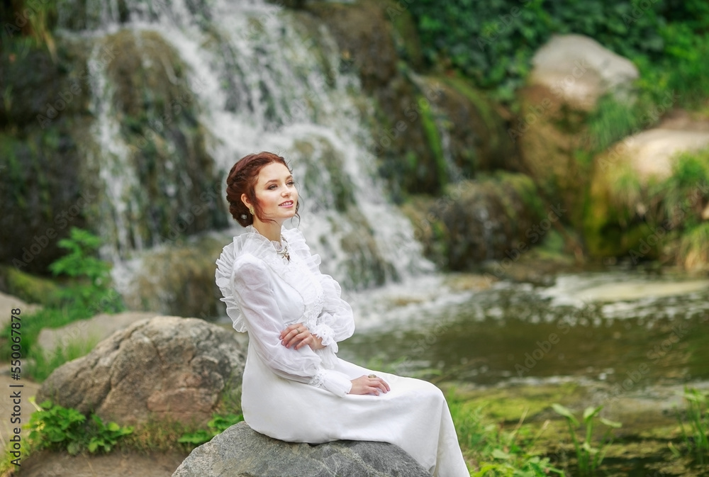 Wall mural Beautiful girl in a vintage long white dress, and a hairstyle in the English style against the backdrop of a waterfall, warm summer