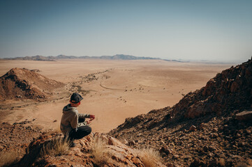 Kleiner Junge sitzt auf einem Felsen unterhalb eines Gipfels am Rande der Aus-Berge und blickt in die Weite der Namib (Klein-Aus Vista, Namibia)