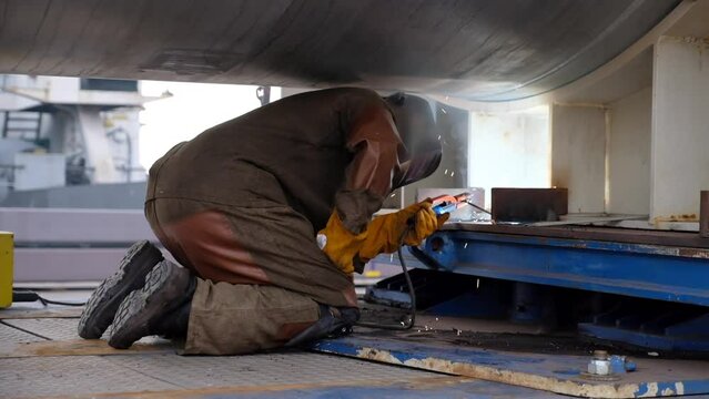 Skilled worker in protective mask welds metal detail kneeling under large turbine. Professional welder in special gloves works in port against drifting ship