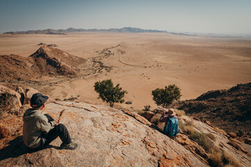 Touristin und ihr Sohn sitzen auf einem Felsen unterhalb eines Gipfels am Rande der Aus-Berge und blicken in die Weite der Namib (Klein-Aus Vista, Namibia)