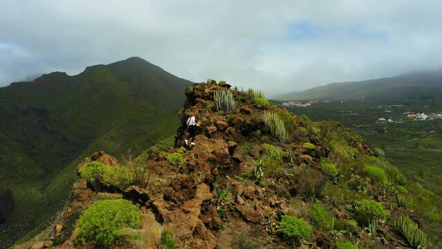 Aerial rear view woman hiking climbs crawling the edge of a cliff in mountain landscape. Tourist enjoys natural volcanic nature and raises her hands up. Teno national park. Tenerife.
