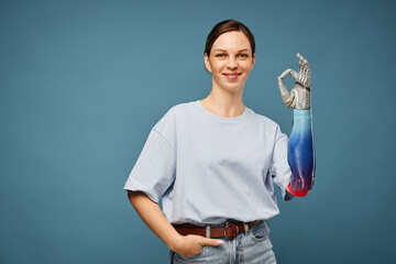 Cheerful young woman with bionic arm showing ok sign, isolated on greyish blu