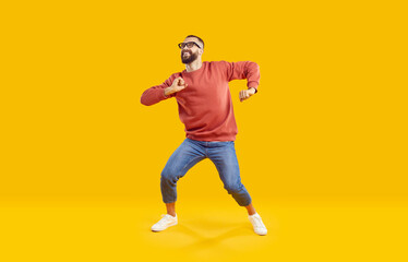 Obraz na płótnie Canvas Cheerful active man enjoying life, rejoicing, dancing and having fun on orange background. Cool stylish Caucasian bearded man in jeans and sweatshirt dancing in good mood. Full length. Web Aner.