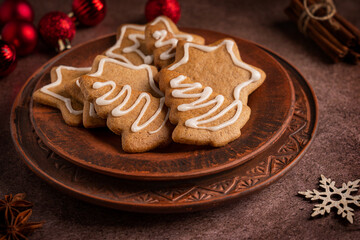 Homemade spiced sweet gingerbread biscuits or crunchy cookies with white sugar icing served on plate on dark brown table with cinnamon, anise and red christmas baubles for new year celebration
