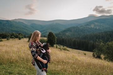 Mom, and child in autumn nature in sunset. Mother with son in sling walk on orange grass in field. Carrying a baby. Holiday trip concept. Family with kid hiking in a mountains. World Tourism Day.