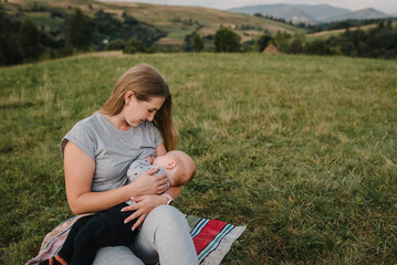 Mother feeding breast newborn baby boy sitting on blanket on grass in field. Family hiking and feeding infants everywhere outdoors. World Breastfeeding Week. Mom hugs child and travels in mountains.
