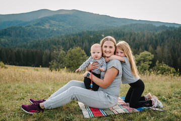 Happy children hugging mom in autumn nature. Young mother with daughter and son sitting on picnic blanket and enjoying the sunset in mountains. Family relaxing and have fun on orange grass in field.