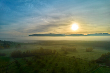 Aerial view of fog landscape at sunrise. Hill range behind agricultural fields in autumn. Blue sky for copy space. Germany, Nurtingen, Tiefenbachtal.