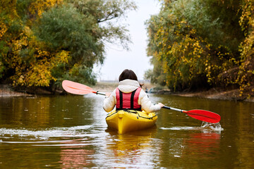 Woman paddle yellow kayak in early autumn at river. Kayaking near overgrown river shore of trees....