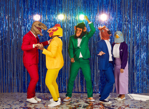Full-size photo of dancing multinational people dressed in bright costumes and rubber masks of various animals. Group of showmen fool around and dance in a club on a shiny blue background and confetti