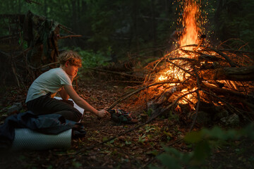 Young caucasian teenage boy adding dry wood to big campfire in the evening time. Camping, traveling and happy teenhood concept image. - obrazy, fototapety, plakaty