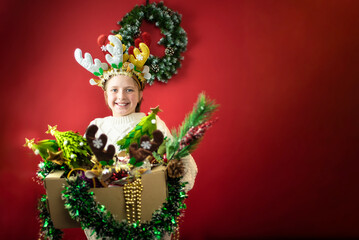 a boy with Christmas deer antlers on his head stands in front of a red wall with a festive wreath.holds three toy Christmas trees	