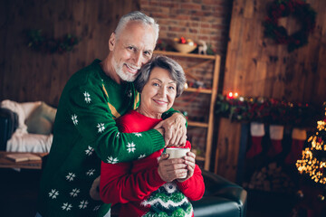 Photo of positive cheerful couple hugs old age seniors staying home wear comfortable sweaters hold cup sweat drink hot cacao indoors xmas atmosphere