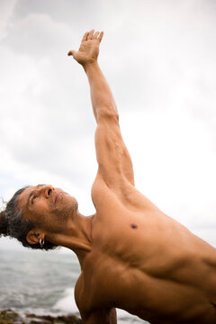 A fit man reaches to the sky while practicing yoga on a rocky beach.