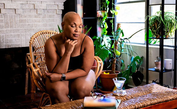 Bald Black woman sitting in wicker chair in living room surrounded by books and plants