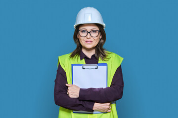 Professional woman in safety vest helmet holding clipboard on blue background