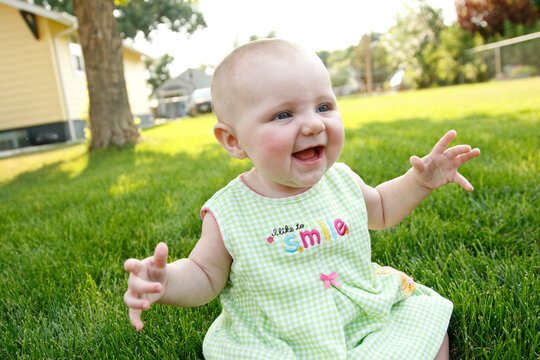 A baby girl in a summer dress smiles and laughs as she sits in the green grass of her backyard in Montana.