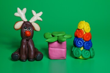 A toy deer with a Christmas tree and a gift made of plasticine on a green background. Christmas decorations.
