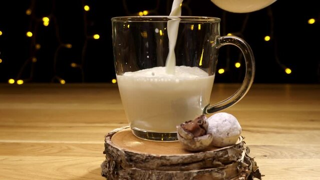 Milk is poured into a transparent glass. Aside some chocolate truffles on a wooden coaster. Dark background with light decoration