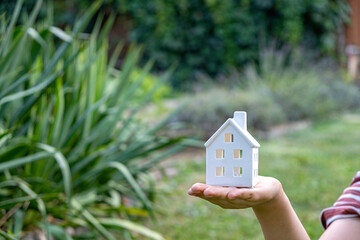 Child's hand holding small toy house on natural green blur background.