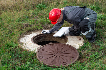 A man working plumber in overalls bent over a water well fixes the measurements made and the...