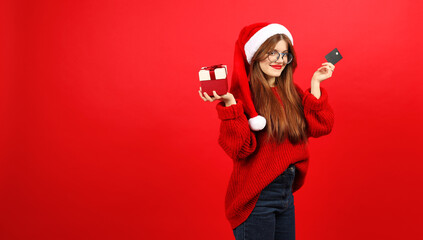 A girl in a Santa hat with a gift and a credit card in her hands on a red background.