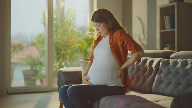 Beautiful pregnant woman is having pain in her stomach. Crying while touching her belly sitting on sofa at home