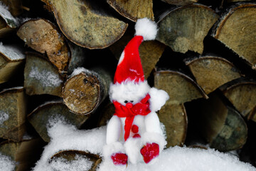 A toy snowman with a red hat sits in the snow near a pile of firewood