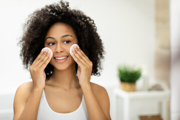 Beautiful smiling black woman cleans her skin with cotton pads and looks in the mirror