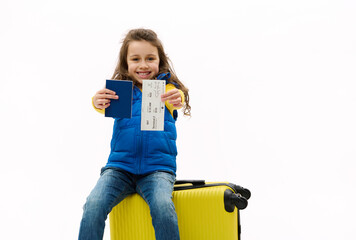 Happy Caucasian little child girl traveler sits on a yellow suitcase, handing a passport and air...