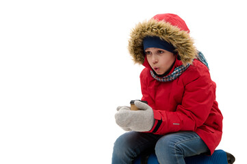 Charming preteen traveler boy in red down jacket and wool mittens, going for vacations, warms his hands with cup of hot drink, sits on suitcase, waits to board a flight. Travelling at winter holidays