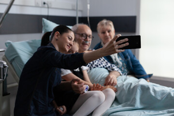 Accompanying hospitalized elderly man for routine checkups capturing cell phone snapshots at geriatric medical tower. Young woman taking selfie with sick father in clinic rest room.