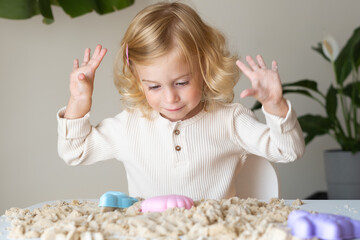 Cute happy caucasian,blonde,curly-haired toddler,baby girl playing with kinetic sand...