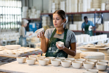 Caucasian young woman potter evaluating quality of new crafted ceramic bowls in workshop.